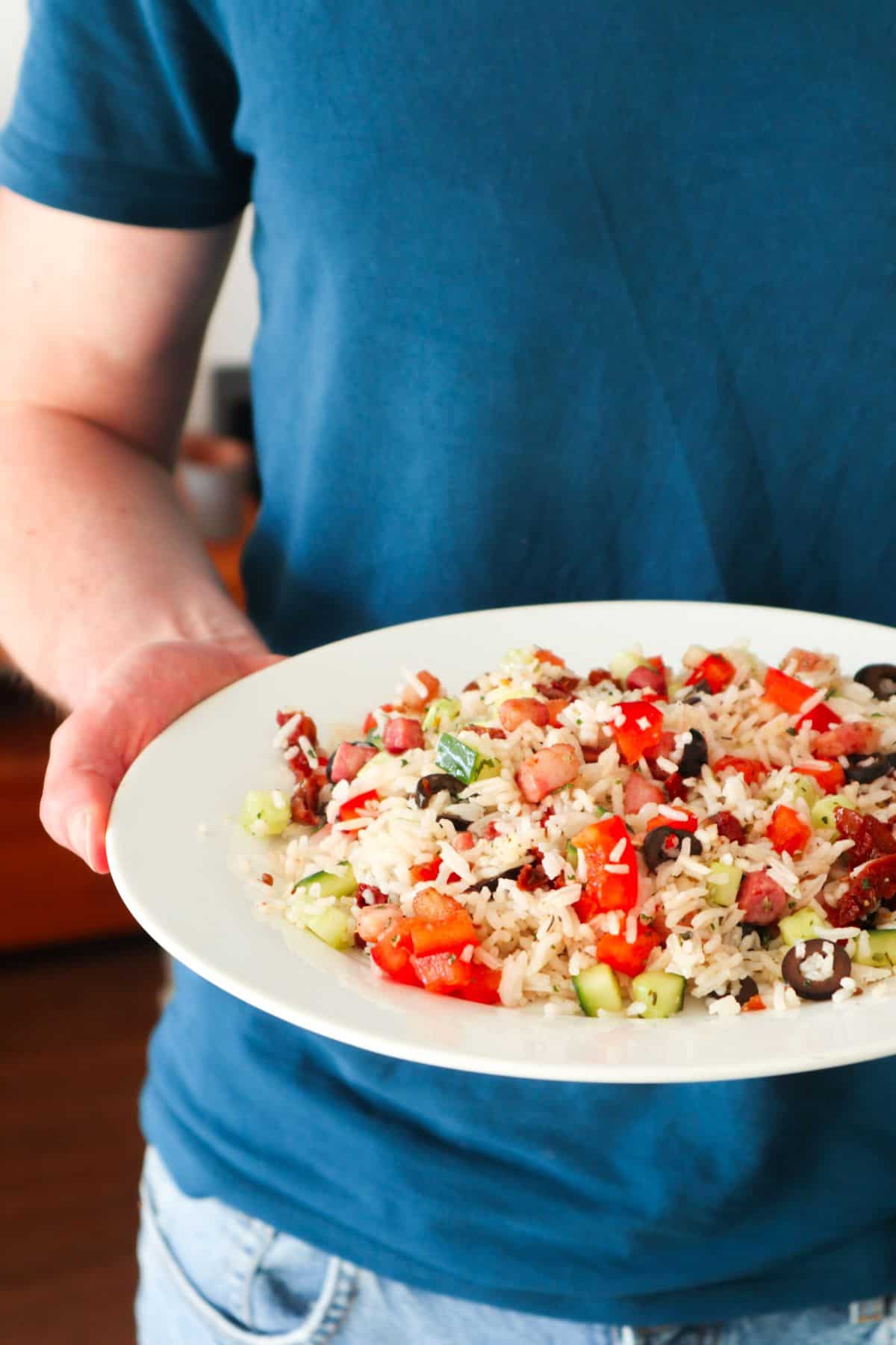 Rijstsalade met spekjes en komkommer op een bord gedragen door een man met een blauw shirt