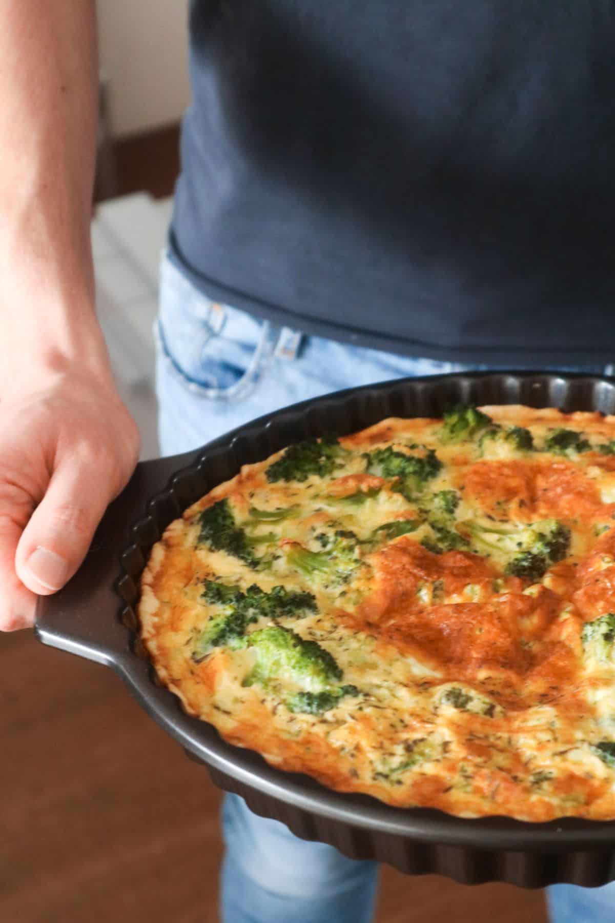 een zelfgemaakte broccoli quiche met spekjes en kaas in een zwarte quichevorm gedragen door een man